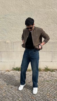 Άνδρας με γυαλιά ηλίου, καφέ μπουφάν, μαύρο πουκάμισο, μπλε τζιν και λευκά αθλητικά παπούτσια στέκεται σε λιθόστρωτο δάπεδο μπροστά σε μπεζ τοίχο.