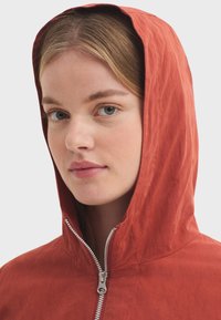 Veste rouge à capuche avec une fermeture éclair frontale, présentant une texture lisse et une coupe décontractée. La capuche encadre le visage tout en ajoutant une touche pratique.