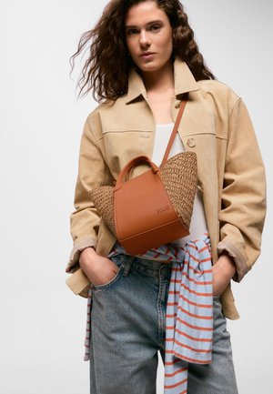 Jeune femme en veste beige et jeans avec chemise rayée nouée à la taille, portant un sac à main marron tissé et en cuir en bandoulière sur la poitrine.