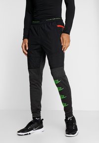 Schwarze und graue Jogginghosen mit strukturierten Einsätzen, einem grünen Logodetail am Bein und einem elastischen Bund mit roten Akzenten.