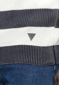 Robno obrobljen pulover z značko logotipa Guess v črno-beli barvi nad modrimi kavbojkami.