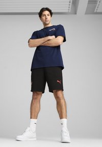 Námořnické bavlněné tričko s nápisem "PORTUGAL", doplněné černými šortkami s červeným logem Puma. Model má na sobě bílé tenisky a sportovní ponožky.
