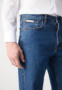 Jean en denim bleu foncé avec une coupe classique, comportant des rivets en cuivre, une fermeture éclair à l'avant et une étiquette Calvin Klein sur la poche arrière.