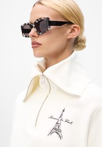 Black and white patterned sunglasses, square shape, with a glossy finish. Model wears a cream zippered top featuring an Eiffel Tower graphic.
