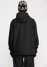 Veste imperméable noire avec capuche, de coupe ample, présentant une texture lisse et un design minimal. Accompagnée d'un bonnet en tricot à motifs.