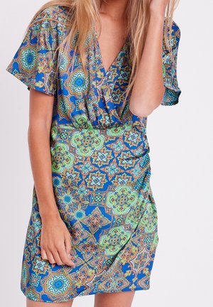 Femme portant une robe bleue avec des motifs floraux et géométriques verts, marron et orange, longueur des manches au-dessus du coude, main reposant près de la hanche.