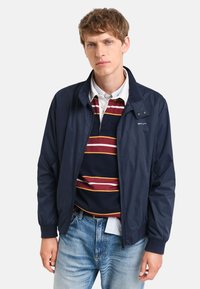 Marineblå nylonjakke med lynlås, ståkrave og sidelommer. Bæres over en stribet polo skjorte, parret med lyseblå jeans.