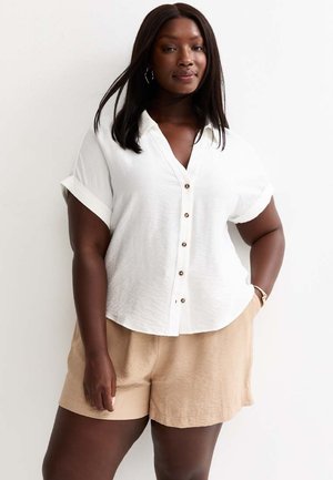 Donna con lunghi capelli scuri che indossa una camicia bianca a maniche corte con bottoni e pantaloncini beige, in piedi con una mano in tasca su sfondo bianco.