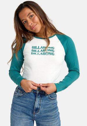 Witte lange mouwen shirt met teal raglanmouwen en "BILLABONG" meerdere keren bedrukt in groen; gecombineerd met high-waisted blauwe jeans.
