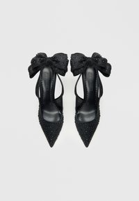 Pantofi negri cu toc înalt și vârf ascuțit, împodobiți cu strasuri și o fundă mare în spate. Textură netedă de piele. Părți laterale deschise.