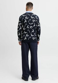 Cardigan en laine bleu marine avec des motifs de rennes et de feuilles blancs. Poignets et ourlet côtelés, associé à un pantalon bleu marine ample.