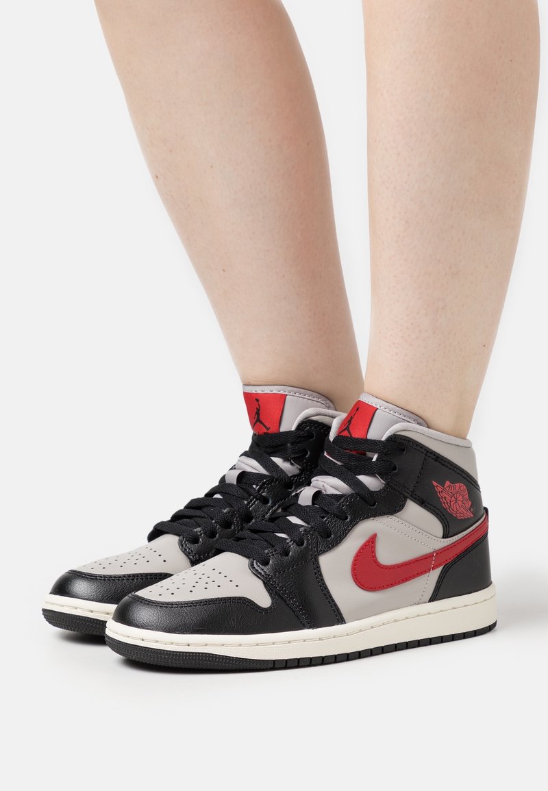 Pieds portant des baskets montantes Nike Air Jordan noires, grises et rouges avec des lacets noirs, debout sur une surface blanche sur fond blanc.