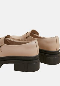 Mocassins synthétiques beiges avec une finition lisse, un bout rond, une sangle décorative et une semelle en caoutchouc noir épaisse.