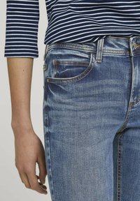 Jean en denim bleu clair avec une coupe droite classique, comportant cinq poches, un léger délavage et des coutures contrastantes. Un haut rayé bleu marine est visible.