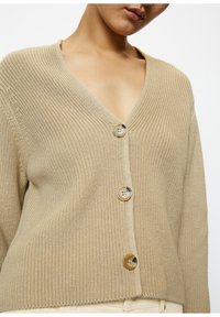 Beige pletený cardigan s výstřihem do V a třemi velkými kulatými knoflíky. Textura je žebrovaná, s volným střihem a minimálními detaily.