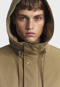 Hnedá kapucňová bunda s prešívanou podšívkou, nastaviteľnými šnúrkami a cvočkovými uzávermi, s hladkým, textúrovaným povrchom.