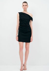 Schwarzes, einseitiges Kleid mit enger Silhouette und leichter Drapierung. Glatte Textur, Oberschenkel-lange, kombiniert mit silbernen Riemchensandalen.