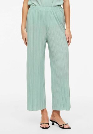 Kvinne iført lysegrønne plissert vidbukser og svarte åpne lavhælte sandaler, stående mot hvit bakgrunn.