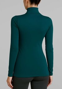 Donna che indossa una maglietta a collo alto fitted di un blu verde scuro e pantaloni scuri, vista di spalle su uno sfondo neutro.