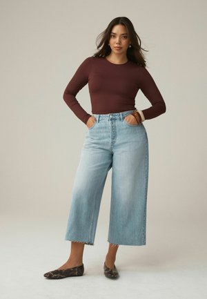 Frau mit langen dunklen Haaren trägt ein bordeauxfarbenes langärmeliges Oberteil, hellblaue, weit geschnittene, verkürzte Jeans, flache Schuhe mit Schlangenmuster und Armbänder, steht mit den Händen in den Taschen.