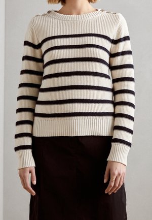 Personne portant un pull crème en tricot avec des rayures horizontales noires et des détails de boutons sur les épaules, associé à une jupe noire.