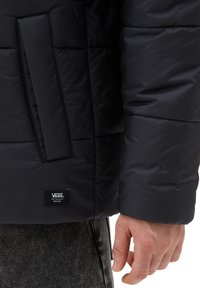 Czarna pikowana kurtka o matowym wykończeniu, z boczną kieszenią i metką z logo Vans na dolnej krawędzi. Teksturowana powierzchnia, luźny krój.