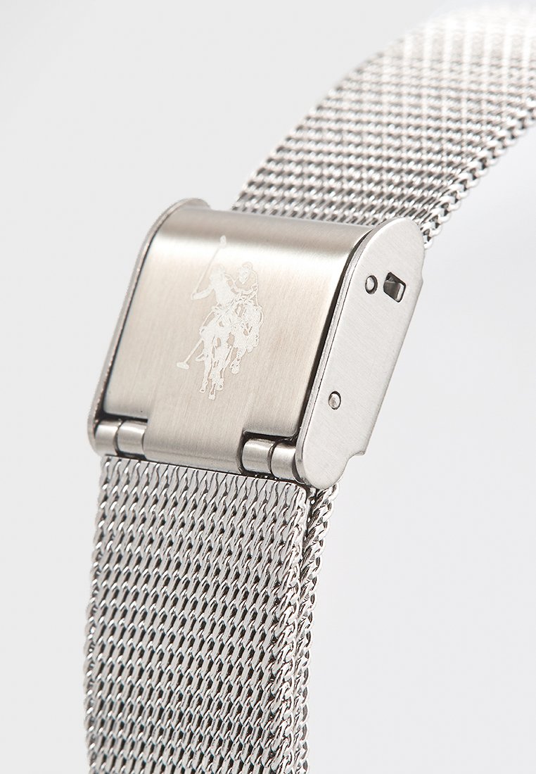 Zilveren mesh armbandhorloge met een metalen sluiting. De sluiting heeft een gegraveerd logo en het mesh heeft een geweven textuur. Minimalistisch ontwerp.