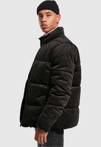 Chaqueta de puffer de pana en gris oscuro con un alto cuello, costuras horizontales y un cierre frontal con cremallera. Textura suave y corte oversize.