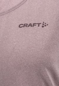 Zbliżenie na fioletową tkaninę w heather, z widocznym dekoltem i ciemnym logo "CRAFT" z okrągłymi kropkami na obszarze klatki piersiowej.