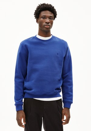 Blauwe sweatshirt met een ronde hals, ribbelmanchetten en -zoom. Heeft een klein embleem op de borst. Gekocht over een wit shirt.