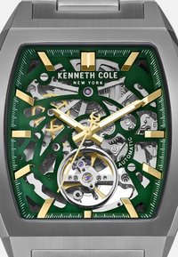Zegarek automatyczny Kenneth Cole posiada zieloną, przezroczystą tarczę, srebrne mechanizmy, złote akcenty oraz unikalną ośmiokątną kopertę z bransoletą ze stali nierdzewnej.