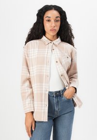 Donna con lunghi capelli ricci che indossa una camicia a quadri beige con bottoni sopra una maglietta bianca e jeans blu, in posa con una mano in tasca.