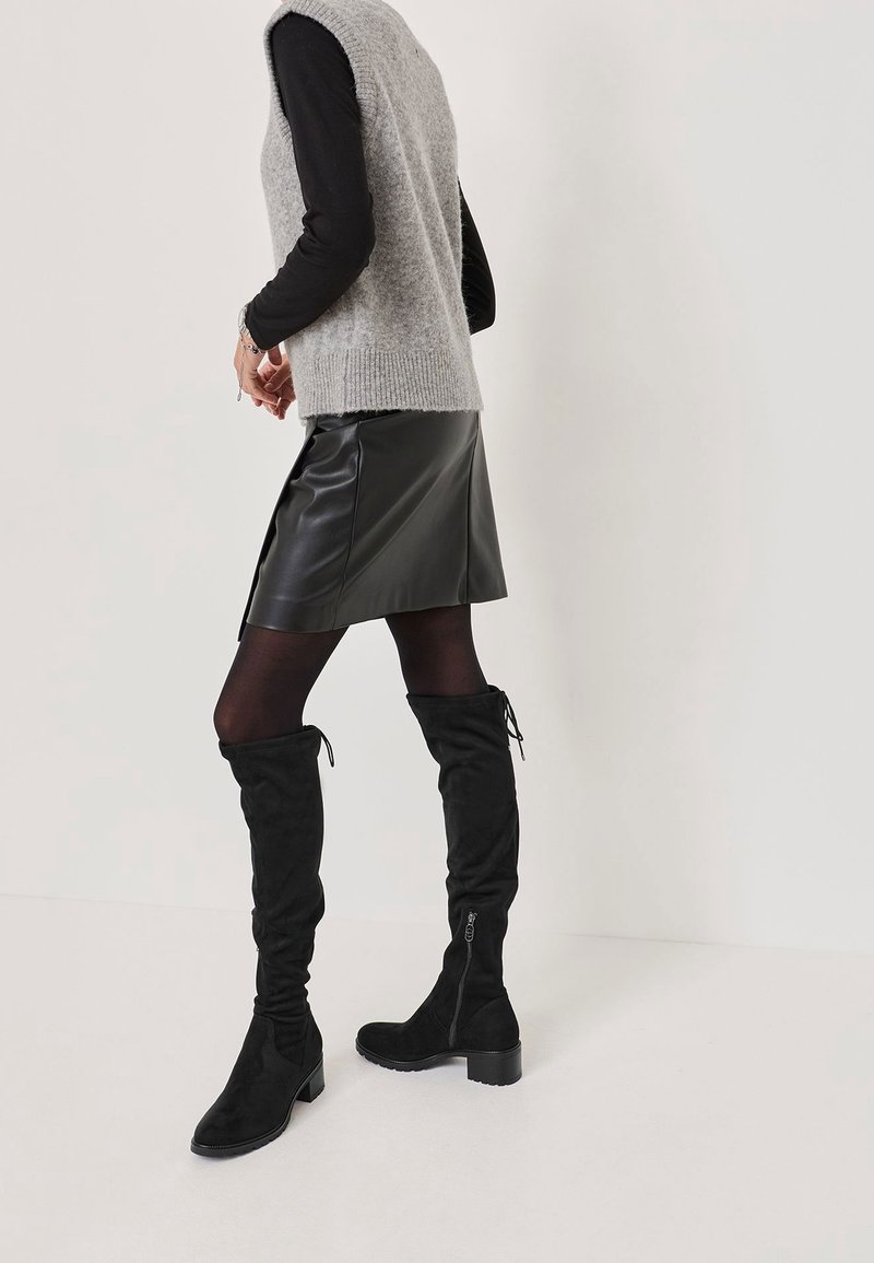 Bottes cuissardes en daim noir à silhouette fine, dotées d'une fermeture éclair latérale et d'une finition texturée. Associées à un pull sans manches gris et une jupe en cuir.