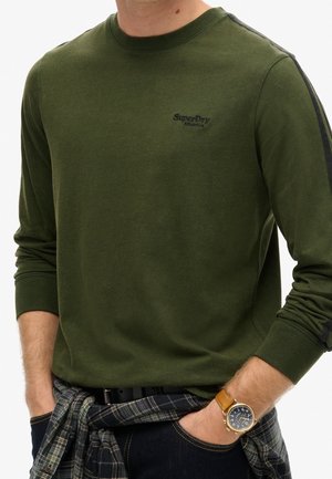 Mężczyzna w oliwkowo-zielonej, długorękawowej koszuli SuperDry z czarnym paskiem na rękawie, koszula w kratę związana wokół talii i brązowy zegarek na skórzanym pasku.