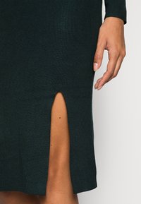 Dunkelgrünes geripptes Kleid mit einem seitlichen Schlitz. Nahaufnahme des Saums und der Hand, die Textur und Passform zeigt.
