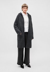Cardigan grigio con texture, dotato di cappuccio e due tasche frontali, dalla vestibilità over size, indossato sopra una camicia bianca e pantaloni neri, abbinato a scarpe nere.