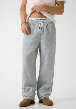 Personne soulève une chemise blanche pour montrer la ceinture blanche du jean Guess, portant un pantalon ample rayé gris clair et des bottes en daim marron.
