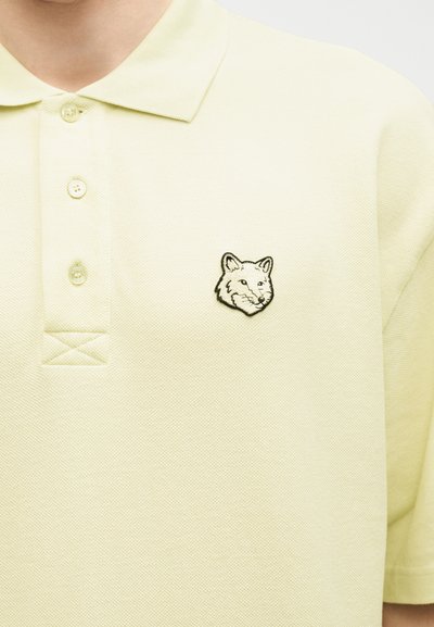 Άτομο που φοράει ανοιχτό κίτρινο πόλο μπλουζάκι με τρία κουμπιά και ένα μικρό ασπρόμαυρο έμβλημα κεφαλής αλεπούς στο στήθος.
