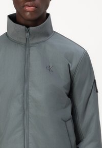 Mężczyzna w szarej kurtce zapinanej na zamek z wysokim kołnierzem i małym logo "cK" na lewej piersi, przedstawiony od ramion do warg.