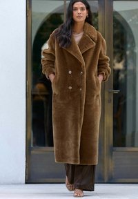 Brun, blød dobbeltradet frakke med brede revers, store knapper og sidelommer; båret over et brunt outfit, foran en glasdør baggrund.