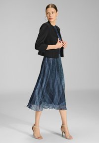 Czarna, krótka kurtka nałożona na niebieską, fakturowaną sukienkę maxi z falistymi wzorami. Ubranie uzupełniają srebrne sandały na obcasie oraz kolczyki w kształcie kół.