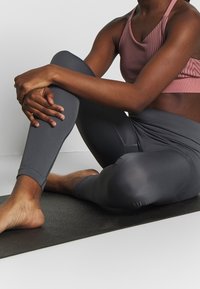 Grå leggings och en rosa randig sport-BH, slät textur, tät passform, person som sitter i lotusställning på en svart yogamatta.