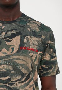 Primer plano de un hombre que lleva una camisa a rayas en verde y beige con el texto rojo "JUST CAVALLI" impreso en el pecho.