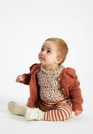 Strikket rustfarvet cardigan over en blomstret skjorte, stribede leggins og hvide sko. Barnet sidder og kigger opad.