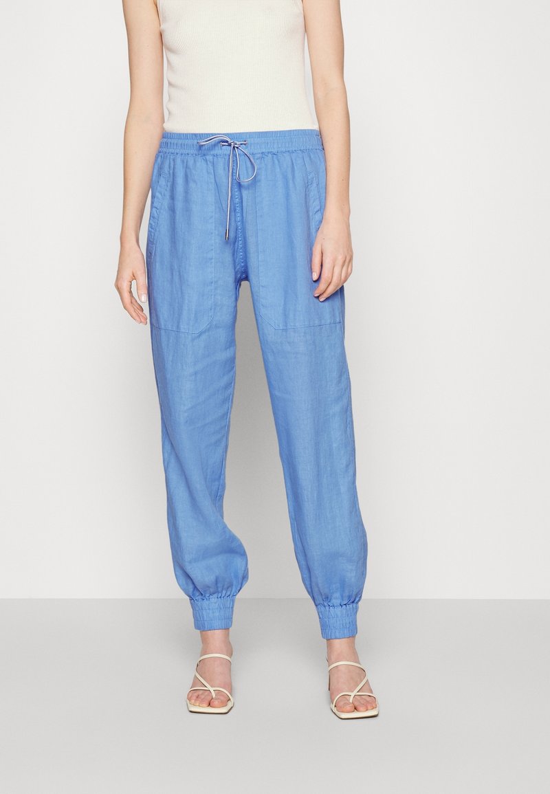 Femme portant un pantalon bleu clair à cordon avec des poignets élastiques et des sandales beiges à brides, debout devant un fond clair uni.