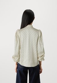 Krémová blúzka s jemným čiernym bodkovaným vzorom, naberanými rukávmi a uzatvoreným golierom, vyrobená z hladkej, ľahkej tkaniny.