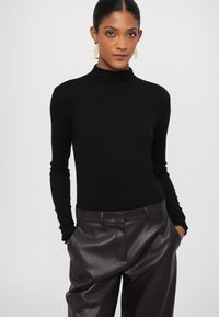 Svart turtleneck genser med lange ermer, kombinert med høyhælte, mørkebrune skinnbukser. Glatt stoffstruktur og tettsittende design.