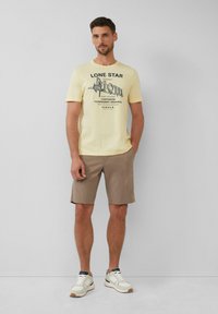 Lichtgele T-shirt met een graphic print met de tekst "LONE STAR" en "MOTEL." Gecombineerd met beige shorts en witte sneakers met grijze accenten.