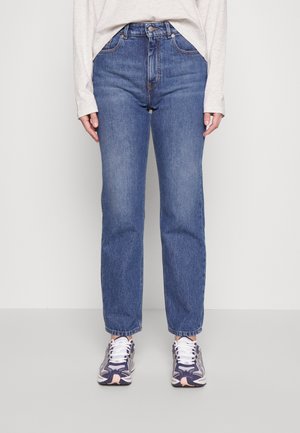 Femme portant un jean bleu taille haute avec un design classique à cinq poches et des baskets blanches avec des accents bleu foncé et rose.