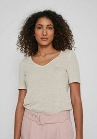 Tricou bej cu decolteu în V, cu mâneci scurte, realizat dintr-un material moale, expus alături de o pereche de pantaloni roz deschis cu talie înaltă, având un accesoriu tip curea.